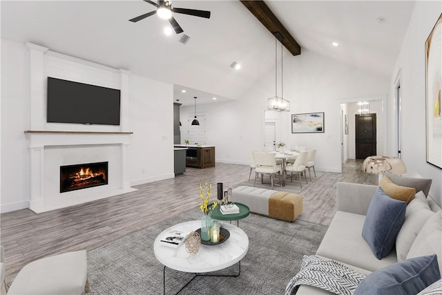 living area featuring high vaulted ceiling, ceiling fan with notable chandelier, a fireplace with flush hearth, light wood-type flooring, and beam ceiling