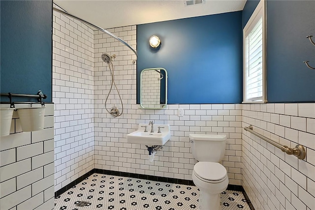 bathroom with tile walls, a tile shower, toilet, a wainscoted wall, and healthy amount of natural light