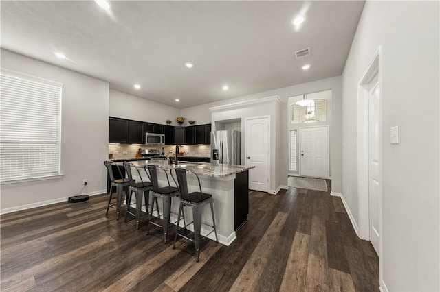 kitchen with dark wood-type flooring, a center island with sink, light stone countertops, appliances with stainless steel finishes, and a kitchen bar