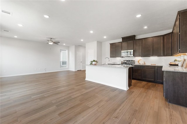 kitchen with light stone countertops, stainless steel appliances, a kitchen island with sink, ceiling fan, and light hardwood / wood-style flooring