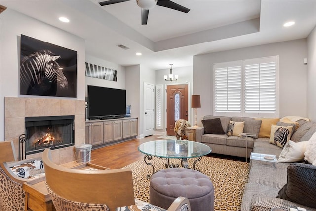 living room with a fireplace, a raised ceiling, ceiling fan with notable chandelier, and light hardwood / wood-style flooring