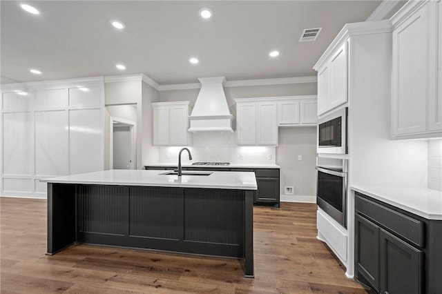 kitchen with stainless steel appliances, custom exhaust hood, white cabinets, and a kitchen island with sink