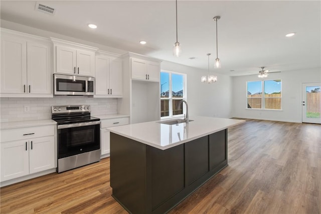 kitchen with appliances with stainless steel finishes, ceiling fan with notable chandelier, white cabinetry, and an island with sink