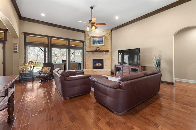 living area with arched walkways, a ceiling fan, crown molding, hardwood / wood-style flooring, and a stone fireplace