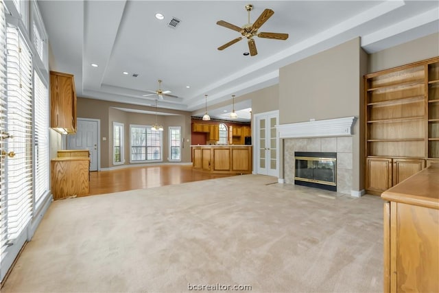 unfurnished living room featuring a raised ceiling, a tile fireplace, ceiling fan, and light colored carpet