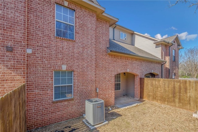 back of house featuring a shingled roof, central AC, fence, and brick siding