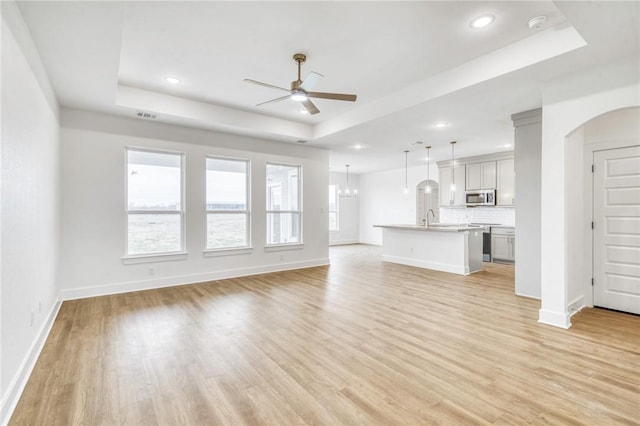 unfurnished living room with arched walkways, a tray ceiling, a ceiling fan, and light wood-style floors