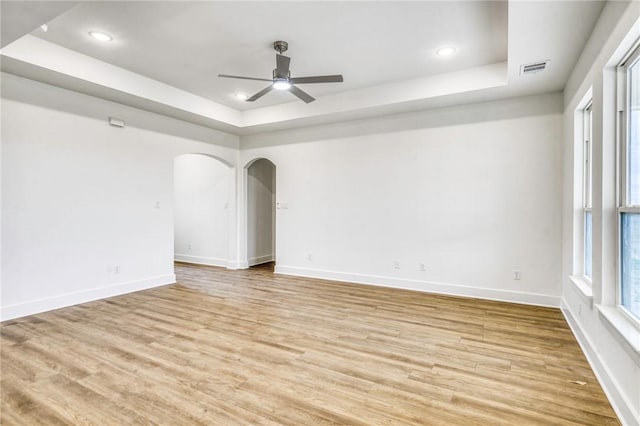 empty room with arched walkways, a raised ceiling, and light wood-style flooring