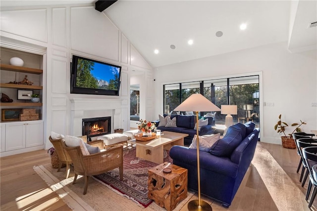 living area featuring high vaulted ceiling, light wood-style flooring, built in features, a lit fireplace, and beamed ceiling