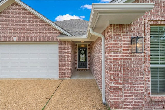view of exterior entry featuring a garage, concrete driveway, roof with shingles, and brick siding