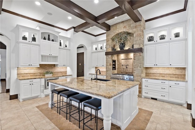 kitchen with beam ceiling, an island with sink, a sink, backsplash, and arched walkways