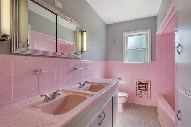 full bath featuring heating unit, tile walls, toilet, a tub to relax in, and double vanity