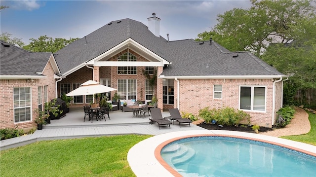 back of house with roof with shingles, brick siding, an outdoor pool, a patio, and a chimney
