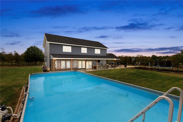 pool at dusk featuring a patio area, a trampoline, an outdoor pool, and a lawn