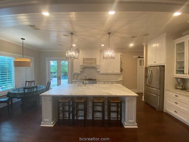 kitchen featuring dark wood-type flooring, decorative light fixtures, stainless steel fridge with ice dispenser, white cabinetry, and an island with sink