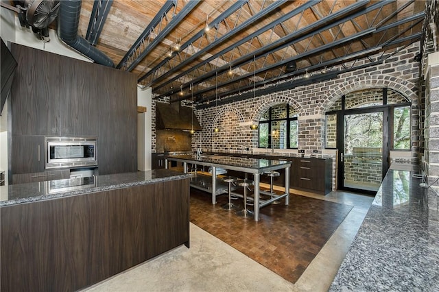 kitchen featuring modern cabinets, stainless steel microwave, dark stone counters, dark brown cabinetry, and brick wall