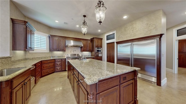 kitchen with light stone countertops, wall chimney exhaust hood, stainless steel appliances, a center island with sink, and hanging light fixtures