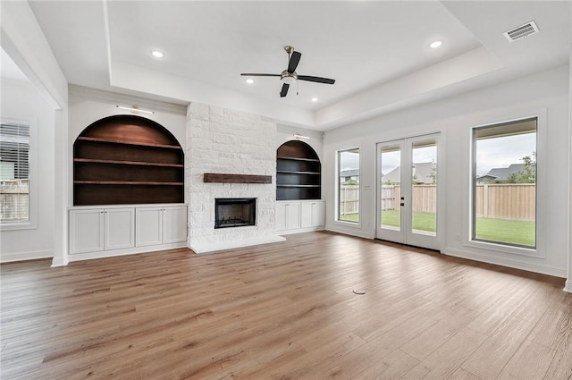 unfurnished living room with a stone fireplace, built in features, a tray ceiling, and light hardwood / wood-style flooring