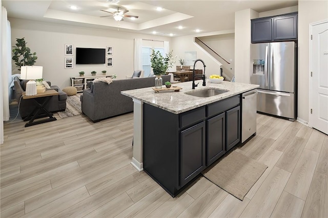 kitchen with sink, light stone counters, a center island with sink, a raised ceiling, and stainless steel appliances