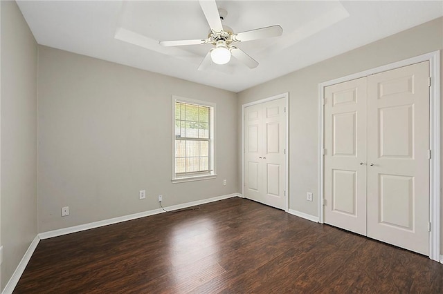 unfurnished bedroom with two closets, ceiling fan, a tray ceiling, and dark hardwood / wood-style floors