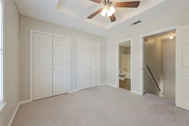 unfurnished bedroom with a raised ceiling, ceiling fan, carpet, and two closets