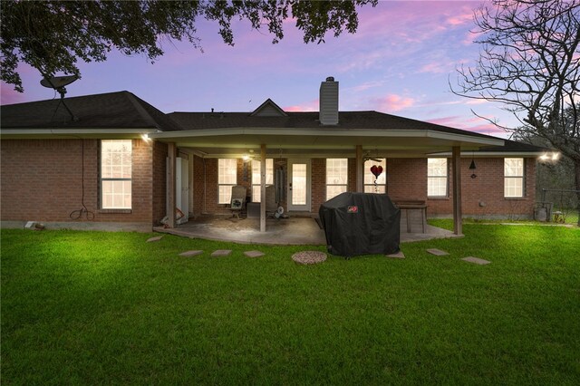 back of property at dusk with ceiling fan, a chimney, a lawn, a patio area, and brick siding