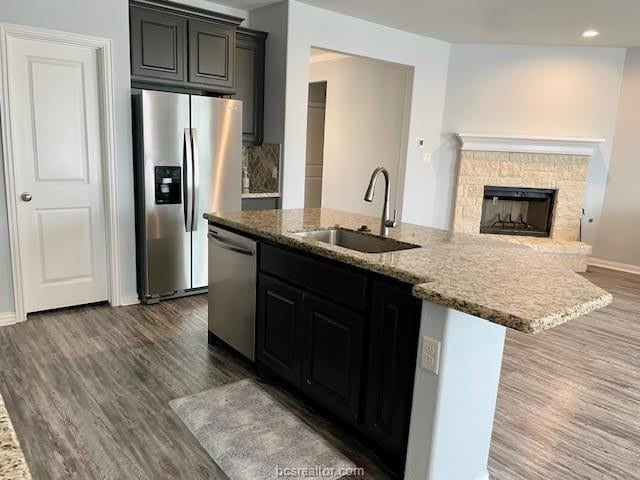 kitchen with a kitchen island with sink, stainless steel appliances, a sink, light stone countertops, and dark wood-style floors