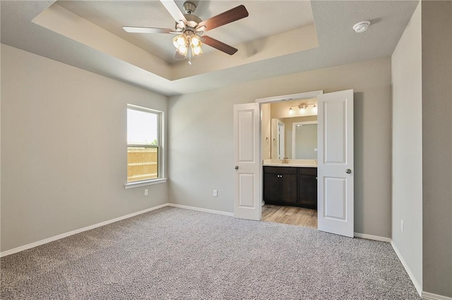 unfurnished bedroom featuring ceiling fan, ensuite bath, a tray ceiling, and light carpet