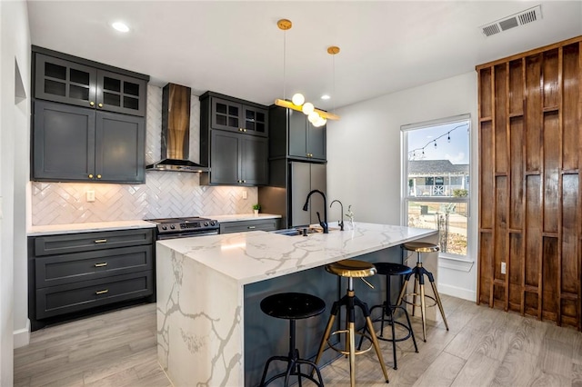 6191 Bethel Cemetery Rd, North Zulch, TX 77872 - kitchen with wall chimney exhaust hood, tasteful backsplash, light wood-style flooring, light stone counters, and a kitchen island with sink