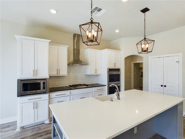 kitchen with an island with sink, wall chimney exhaust hood, arched walkways, and stainless steel appliances