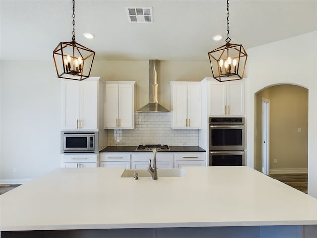 kitchen featuring visible vents, an island with sink, wall chimney exhaust hood, appliances with stainless steel finishes, and pendant lighting