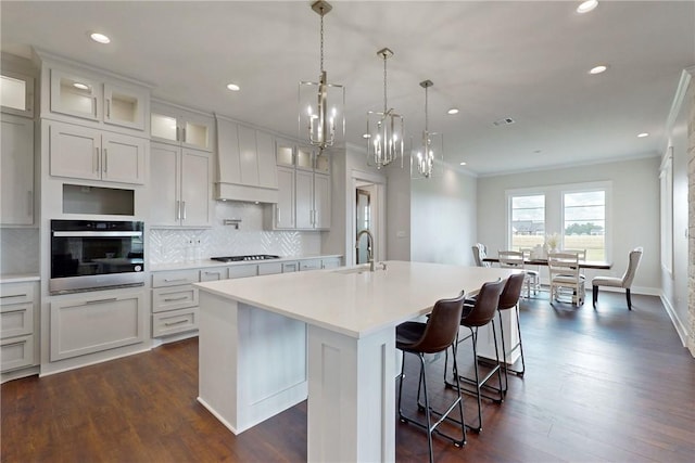 kitchen with stainless steel oven, a kitchen island with sink, sink, tasteful backsplash, and white cabinetry