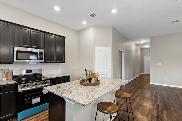 kitchen with light stone counters, dark wood-type flooring, stainless steel appliances, an island with sink, and tasteful backsplash