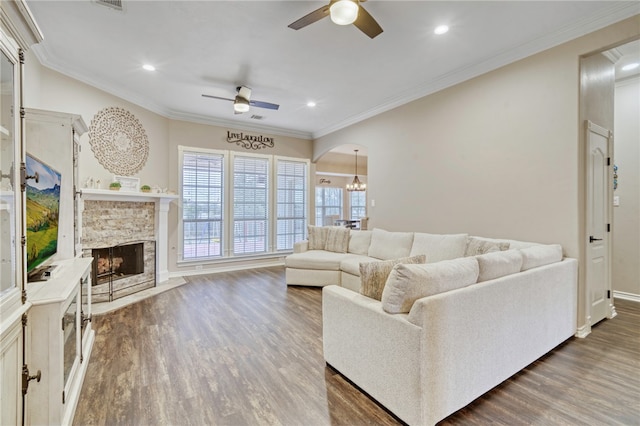 living room with crown molding, a stone fireplace, dark wood-type flooring, and ceiling fan with notable chandelier