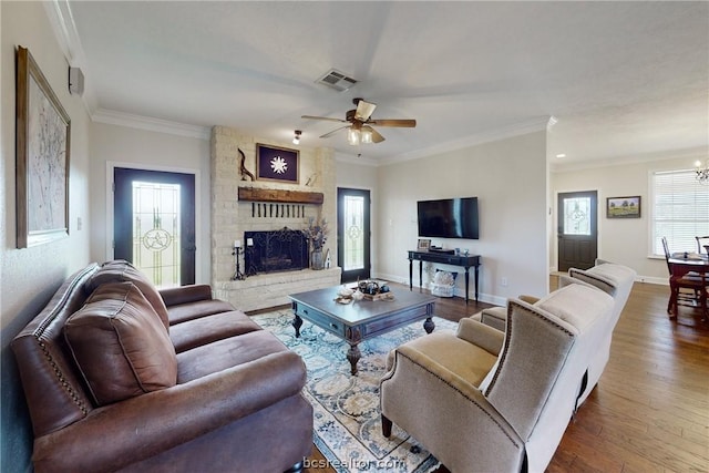 living room with ornamental molding, ceiling fan with notable chandelier, wood-type flooring, a stone fireplace, and plenty of natural light