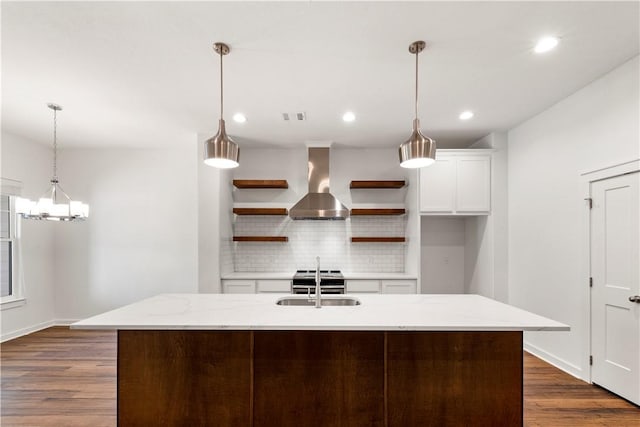 kitchen featuring hanging light fixtures, wall chimney exhaust hood, light stone countertops, an island with sink, and tasteful backsplash