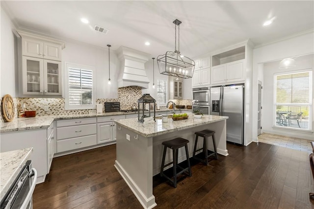 kitchen featuring custom range hood, visible vents, appliances with stainless steel finishes, a kitchen island with sink, and a kitchen bar