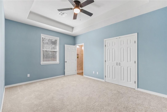 unfurnished bedroom featuring ensuite bathroom, ceiling fan, a tray ceiling, light carpet, and a closet