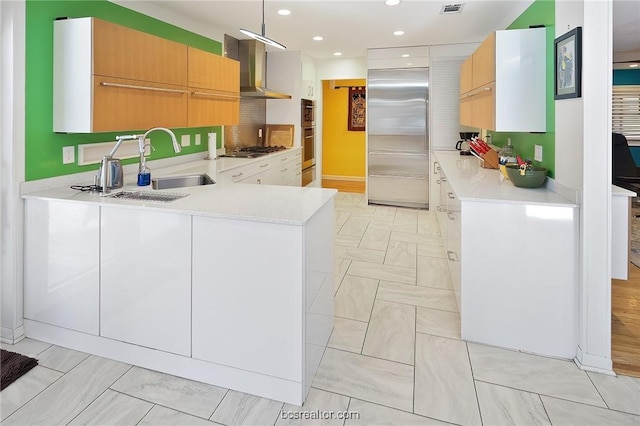 kitchen with kitchen peninsula, appliances with stainless steel finishes, wall chimney exhaust hood, sink, and white cabinetry