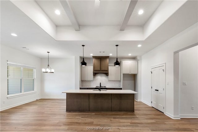 kitchen with tasteful backsplash, beam ceiling, decorative light fixtures, light hardwood / wood-style flooring, and an island with sink