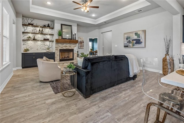 living room featuring light wood-type flooring, a raised ceiling, ceiling fan, crown molding, and a fireplace