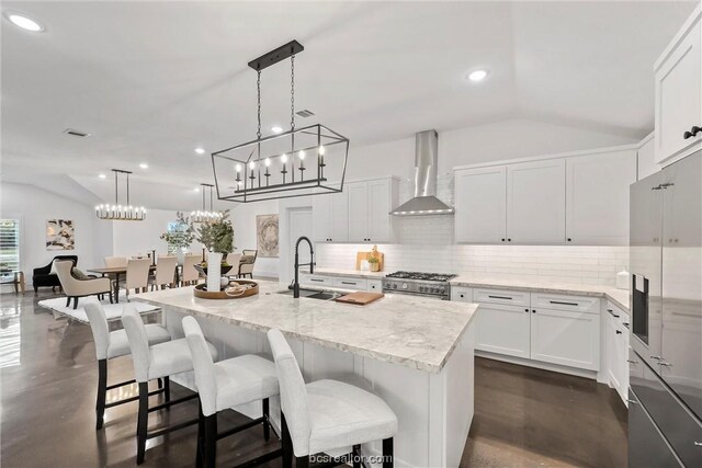 kitchen with pendant lighting, white cabinets, wall chimney exhaust hood, high end stove, and an island with sink