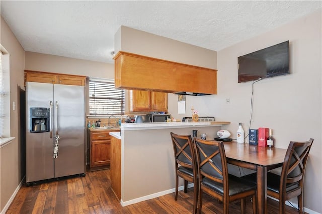 kitchen with brown cabinetry, dark wood-type flooring, a peninsula, light countertops, and stainless steel refrigerator with ice dispenser