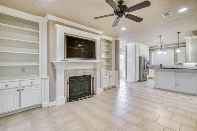 unfurnished living room with built in features, a ceiling fan, visible vents, a tile fireplace, and crown molding