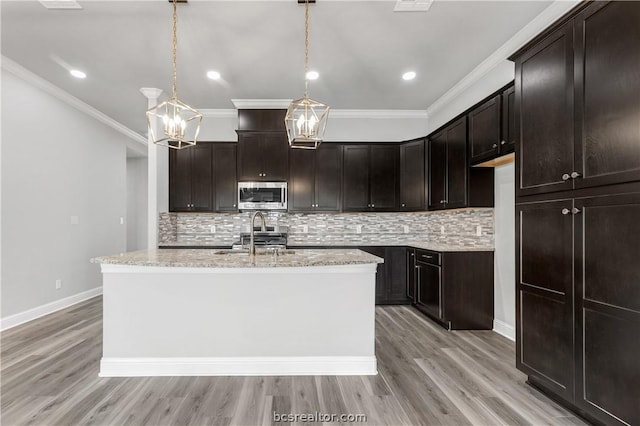 kitchen with hanging light fixtures, ornamental molding, light wood-type flooring, appliances with stainless steel finishes, and an island with sink