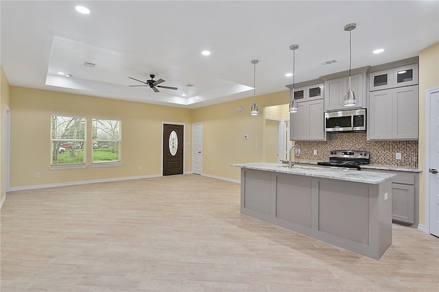 kitchen featuring gray cabinets, hanging light fixtures, stainless steel appliances, a tray ceiling, and an island with sink