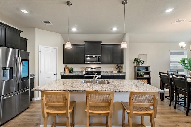 kitchen with stainless steel appliances, a kitchen island with sink, sink, light hardwood / wood-style flooring, and hanging light fixtures