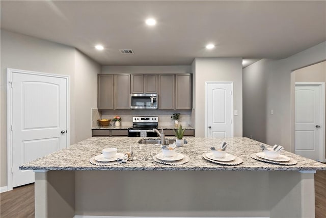 kitchen with a kitchen island with sink, gray cabinetry, dark wood-style flooring, appliances with stainless steel finishes, and light stone countertops