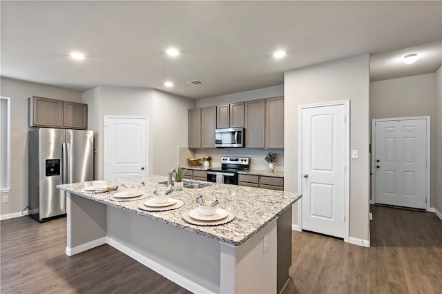 kitchen with light stone counters, stainless steel appliances, a sink, visible vents, and a center island with sink