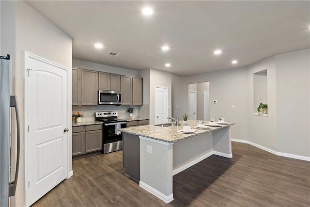 kitchen featuring a center island with sink, stainless steel appliances, gray cabinetry, a sink, and light stone countertops